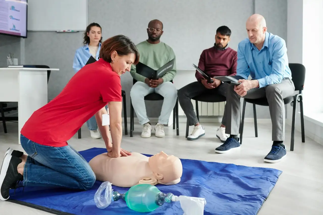 Instructor in red shirt kneeling beside a CPR manikin on a blue mat, class seated behind.