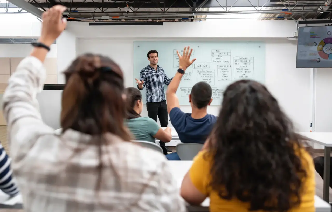 Instructor presenting to a classroom, one attendee raising a hand, whiteboard/flip chart behind.